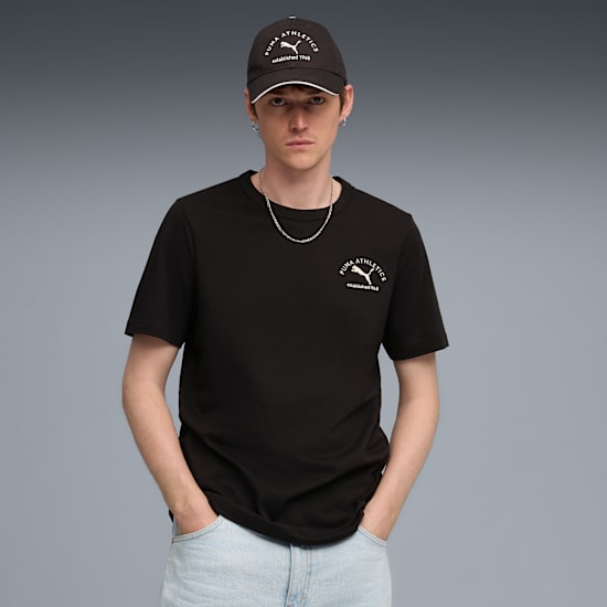 A model with hands in pockets, poses on grey wearing a cap and black t-shirt.