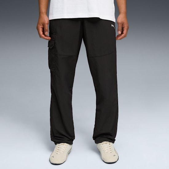 A model stands on a grey backdrop wearing a white t-shirt, black trousers and beige trainers.