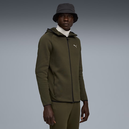 A model stands against a grey backdrop, wearing an olive zip-up hoodie and bucket hat.