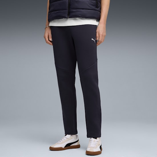 Against grey, a model wears dark trousers, trainers, vest and a white t-shirt.