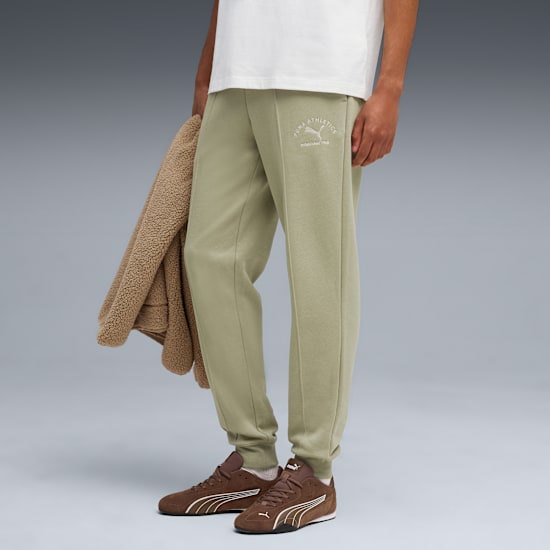 Su sfondo grigio, un modello indossa pantaloni da jogging verde chiaro e scarpe da ginnastica marroni, tenendo una giacca.