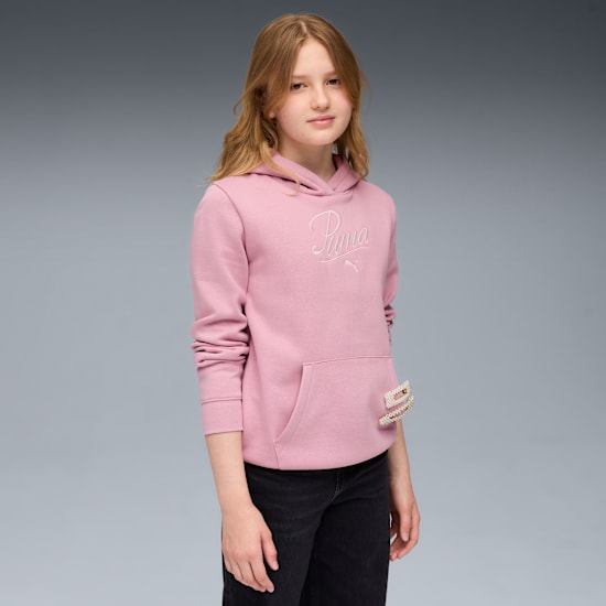 Een model staat tegen grijs gekleed in een roze hoodie met zwarte jeans.