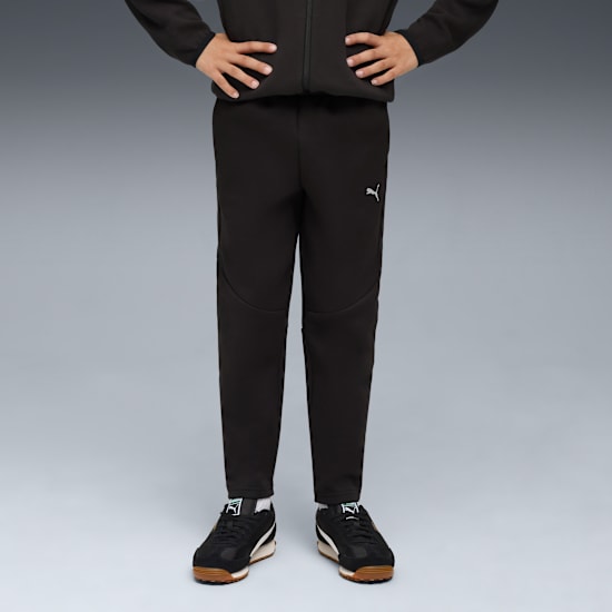 Against grey, a model poses in black Puma tracksuit bottoms and trainers, hands on hips.