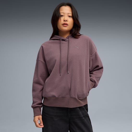 Against grey, a model poses in a mauve hoodie and dark trousers.