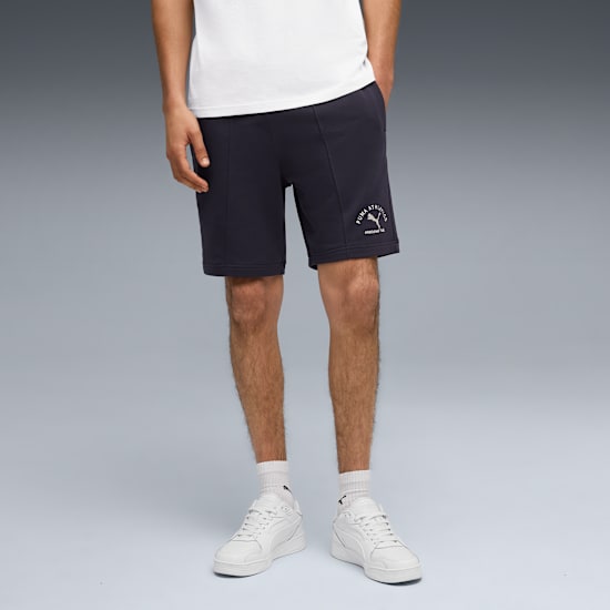 Against grey, a model wears a white tee, navy shorts with logo, white socks and trainers.