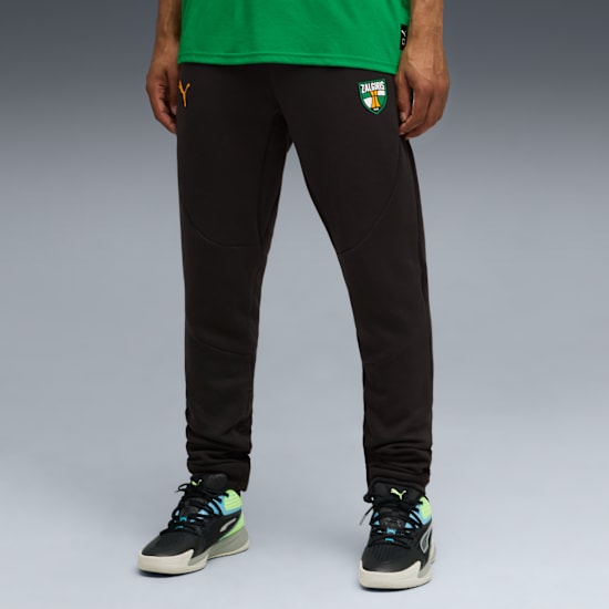 Against a grey backdrop, a model wears a green t-shirt, dark trousers, and trainers.