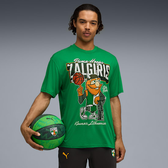 Een model houdt een basketbal vast, draagt een groen T-shirt met een cartoon op een grijze achtergrond.