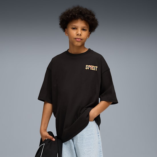 Ein Jugendlicher posiert vor grauem Hintergrund, trägt ein schwarzes T-Shirt mit 'SPIRIT'-Text und hellblaue Jeansshorts.