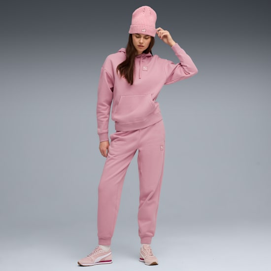 A model in front of grey wears a pink hat, hoodie, joggers and trainers.