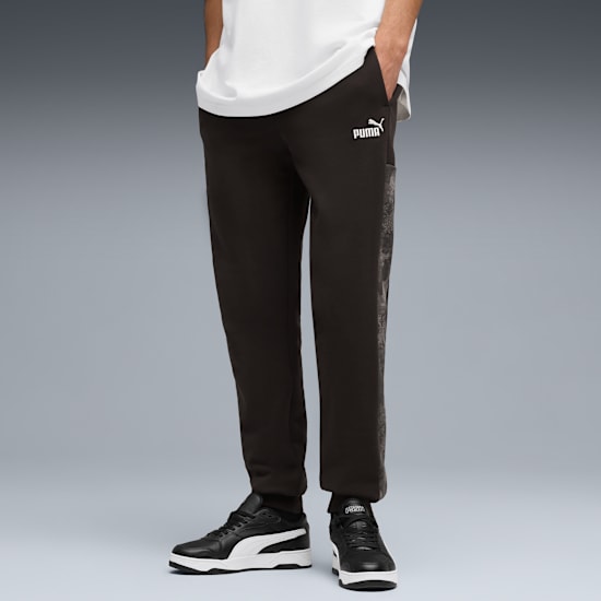 Against grey, a model wears a white tee, dark trousers with a Puma logo, and black trainers.