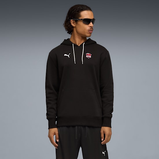 A model with sunglasses poses in front of a grey backdrop wearing a black hoodie and trousers.