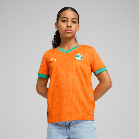 Against grey, a model poses wearing an orange patterned t-shirt with green trim and light blue denim shorts.