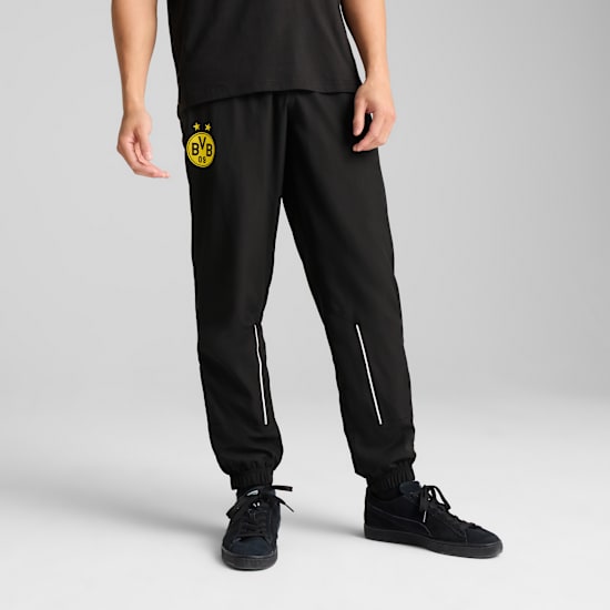 Sur un fond gris, un modèle pose en jogging noir avec un logo jaune et des baskets noires.