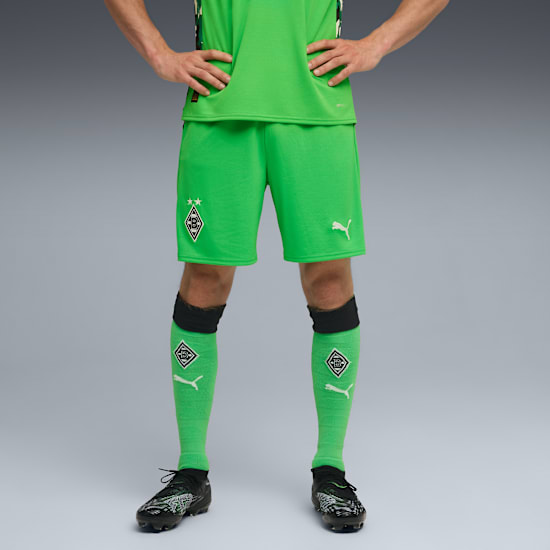 Against grey, a model poses in green soccer attire and black shoes, hands on hips.