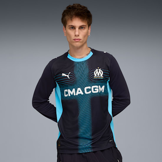 Against grey, a model poses wearing a long-sleeved black and blue sports shirt.