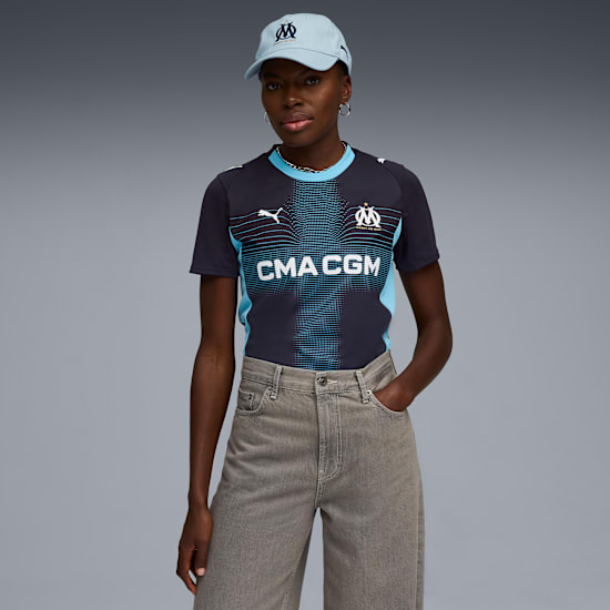 A model poses against grey wearing a cap, navy shirt and grey trousers.