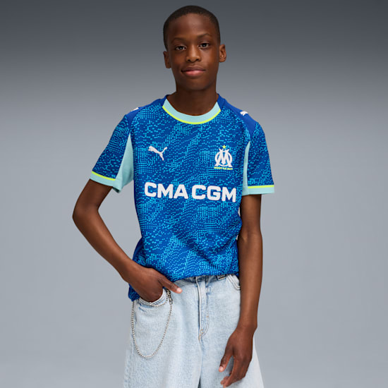 A model poses against a grey backdrop, wearing a blue football shirt and light denim shorts.