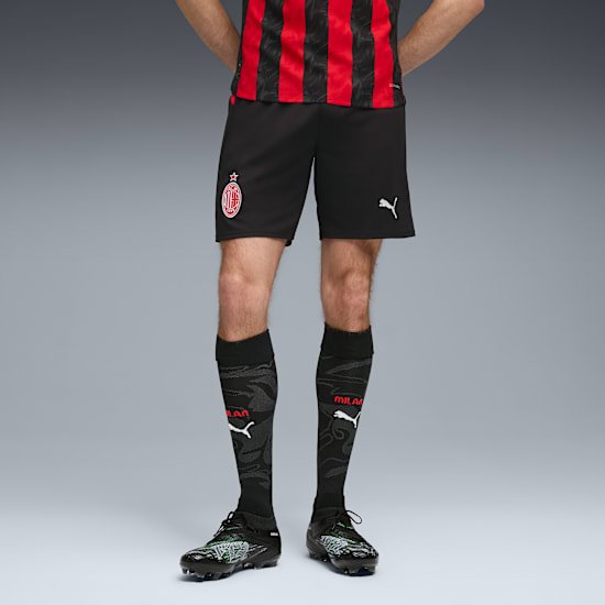 Against grey, a model wears a red and black top with black shorts, socks and football shoes.