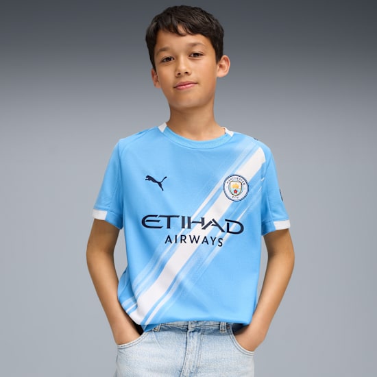 A child poses against grey in a light blue football shirt and light blue jeans.