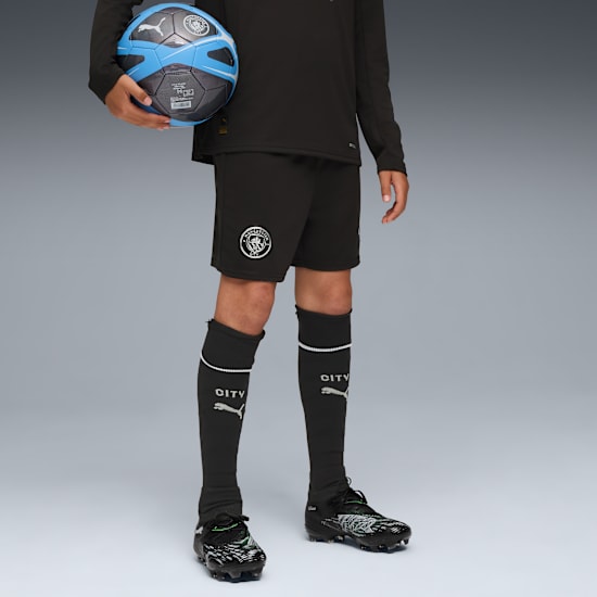 Against grey, a model holds a football, wearing black kit, socks and football boots.