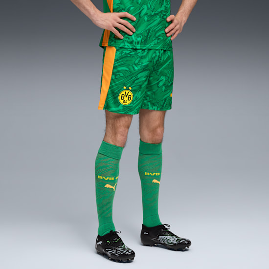 A model poses against grey, wearing green and yellow BVB football shorts, socks and black football boots.
