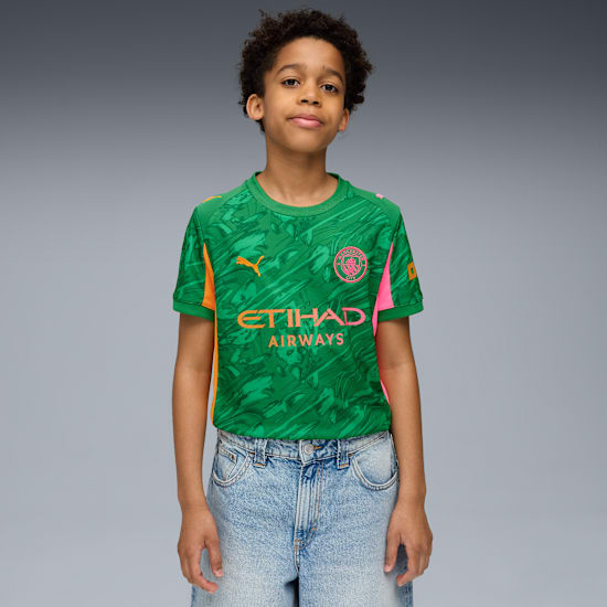 A child poses against grey, wearing a green patterned t-shirt and light blue denim shorts.