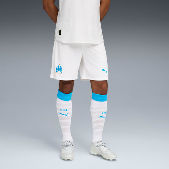 Against grey, a model wears a white Marseille kit with blue accents and white footwear.