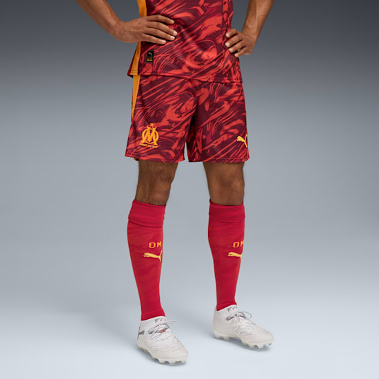 A model displays a red patterned kit with matching socks and white football boots on grey.