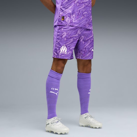 Against grey, a model wears lilac patterned shorts, top, socks and white football boots.