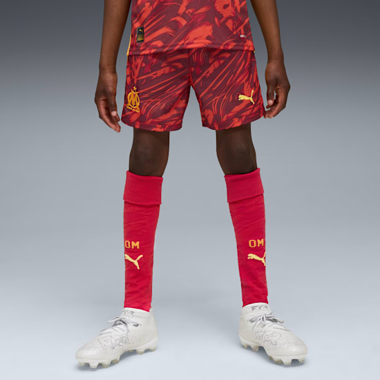 A model stands on a grey backdrop, wearing red patterned shorts, socks and white football boots.