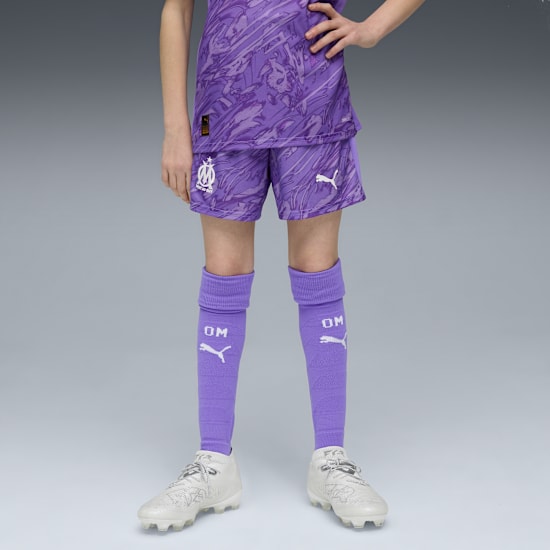 A model in front of a grey background wearing a purple football kit and white football boots.
