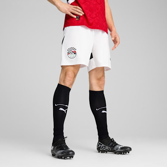 Een model draagt een rood shirt, witte short, zwarte sokken en schoenen tegen een grijze achtergrond.