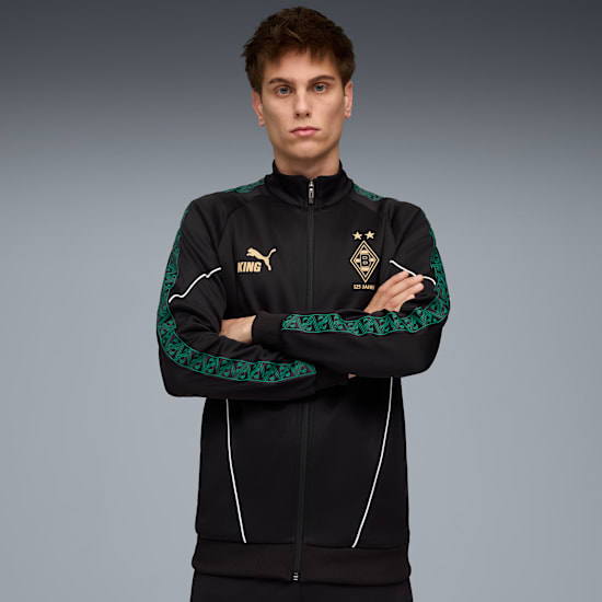 Model pozuje ze skrzyżowanymi rękami, ubrany w czarno-zieloną kurtkę na szarym tle.