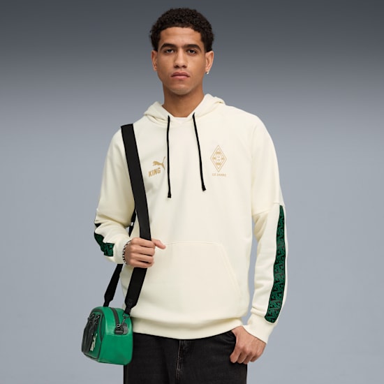 Model na szarym tle ma na sobie kremową bluzę z kapturem, czarne dżinsy i zieloną torbę.