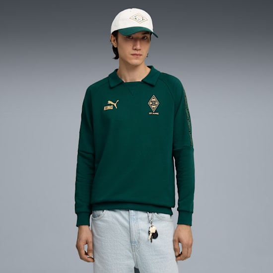 Model pozuje na szarym tle, ubrany w zieloną bluzę, jasne dżinsy i czapkę.