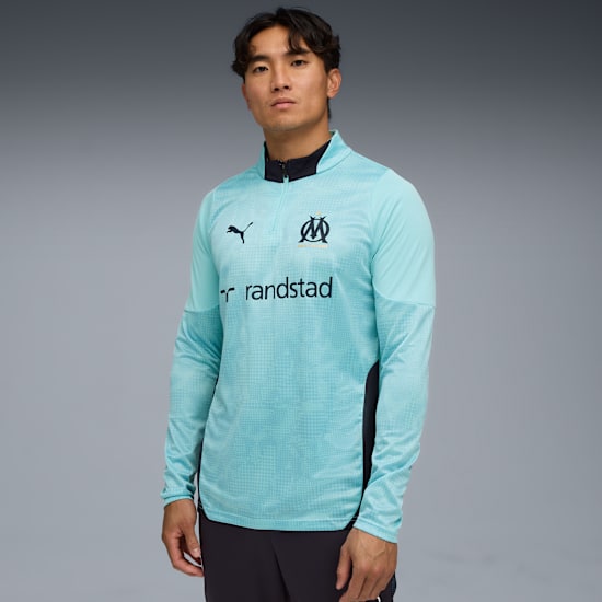 Ein Model vor grauem Hintergrund, trägt ein hellblaues Langarmshirt und dunkle Hose.