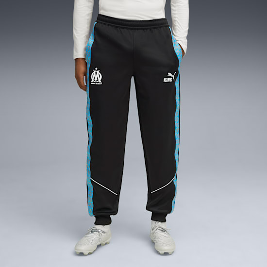 Op grijs draagt model zwarte trainingsbroek met blauwe strepen en witte voetbalschoenen.