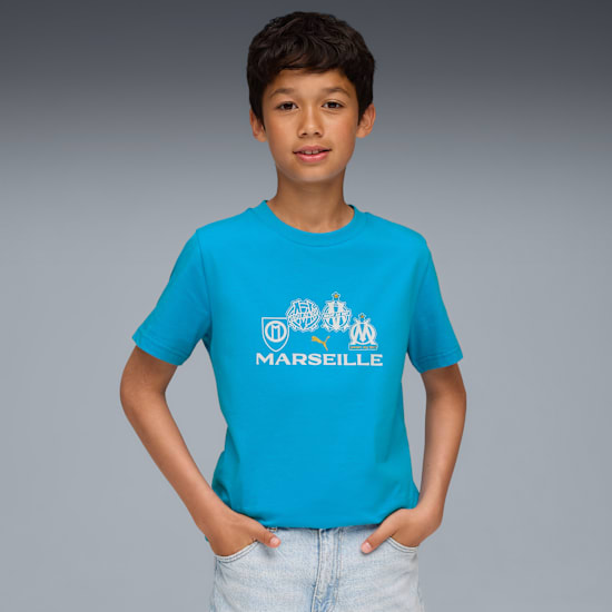 Vor grauem Hintergrund posiert ein Kind in blauem T-Shirt mit weißen Marseille-Emblemen.