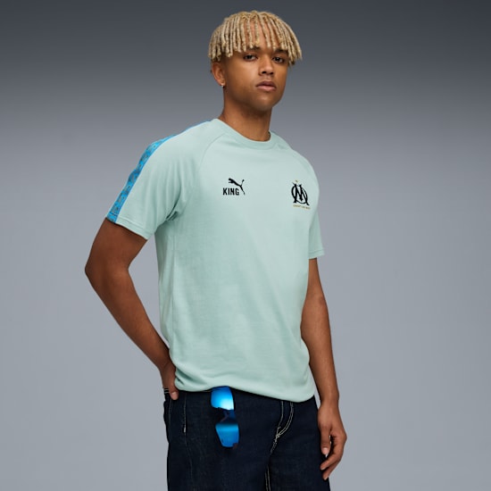 Un modello posa su sfondo grigio, indossa una t-shirt verde chiaro e jeans scuri, con la mano in tasca.