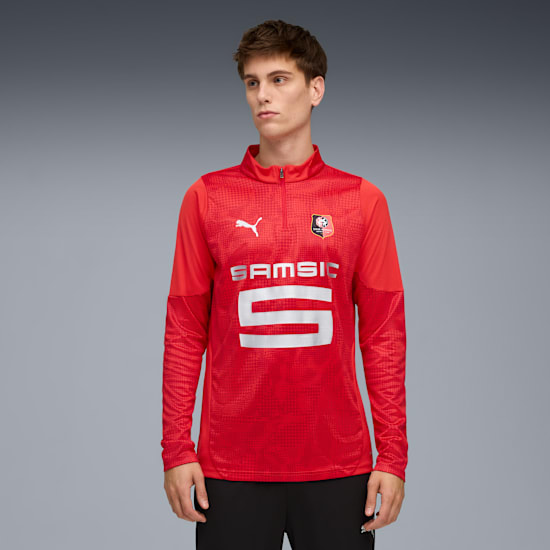 Vor grauem Hintergrund trägt ein Model ein rotes Langarmshirt, schwarze Hose und blickt zur Seite.