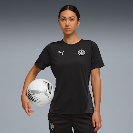 Against grey, a model holds a football, wearing a black t-shirt and shorts with logos.