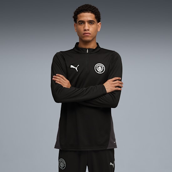 Against grey, a model with crossed arms wears a black Puma Manchester City tracksuit.