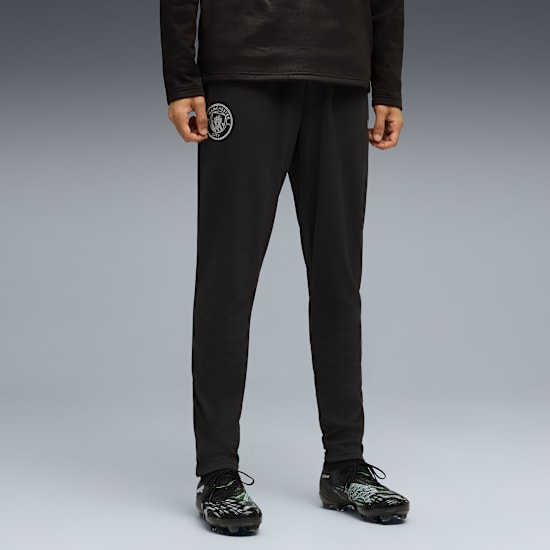 Against grey, a model wears black tracksuit bottoms with a team logo and football boots.