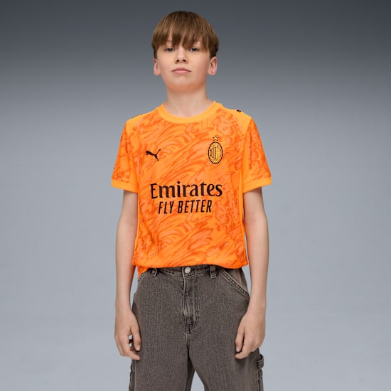 A young model stands against a grey backdrop, wearing an orange patterned t-shirt and grey jeans.