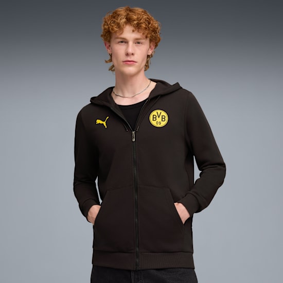 Model pozuje na szarym tle, ubrany w czarną bluzę z kapturem z żółtymi logotypami.