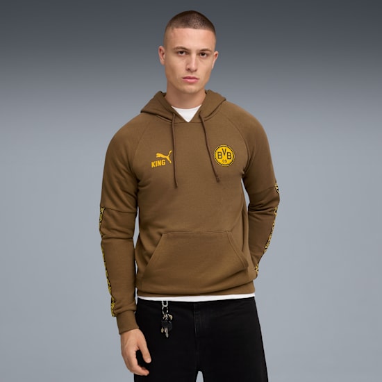 A model stands against grey, wearing a brown hoodie with yellow logos and black jeans.