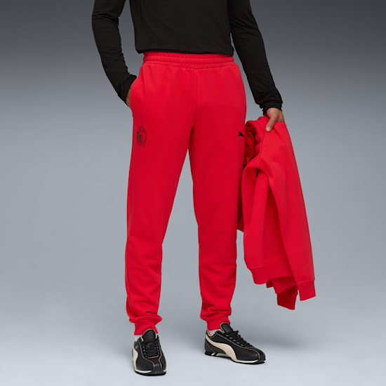 Against grey, a model poses wearing red joggers, a black top, and trainers, holding a red jacket.
