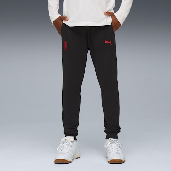 Sur fond gris, un modèle porte un pantalon noir à emblèmes rouges et des baskets blanches.