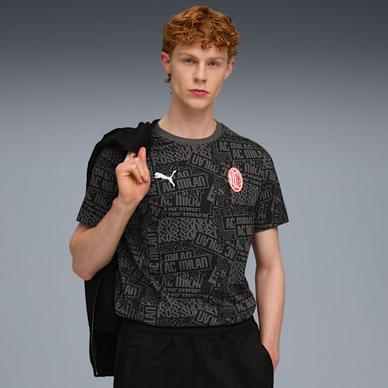 Against a grey background, a model poses in a patterned t-shirt and dark trousers.