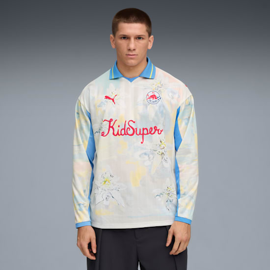 Against grey, a model wears a floral long-sleeved shirt, dark trousers, looking forward.